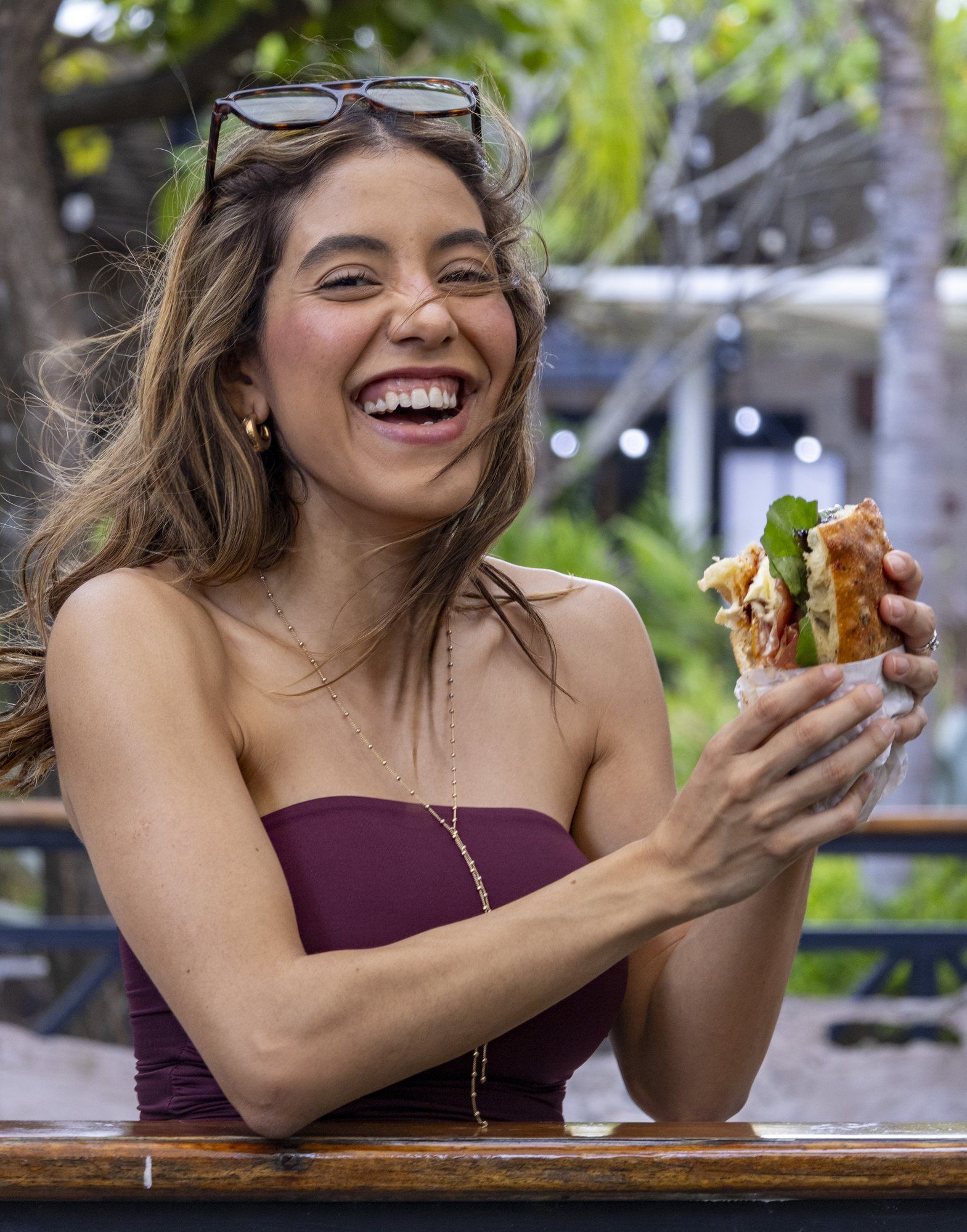 A woman laughing at an outdoor table, holding a focaccia sandwich